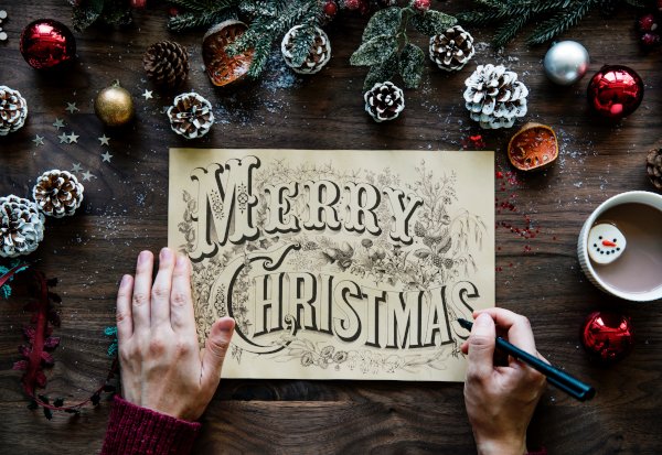 2 hands holding a 'Merry Christmas' sign in fancy calligraphy, one hand still holding a pen, assorted christmas decorations on a table.